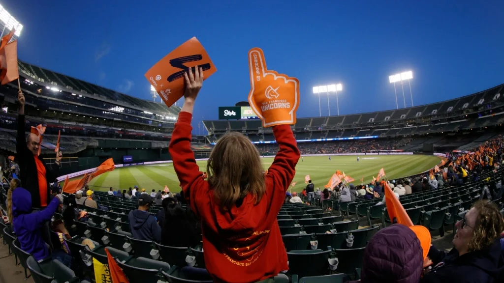 San Francisco Unicorns cricket fans cheering with foam fingers and team flags at Oakland Coliseum during a Major League Cricket (MLC) match in the evening.