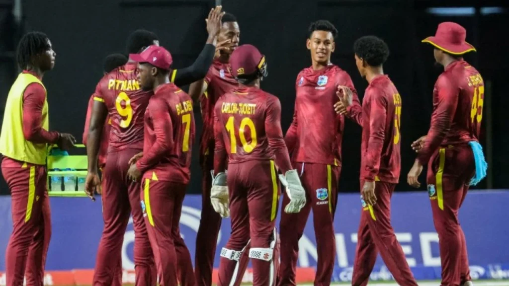 West Indies Under-19 cricket team players in maroon jerseys celebrating on the field during a match under stadium lights.